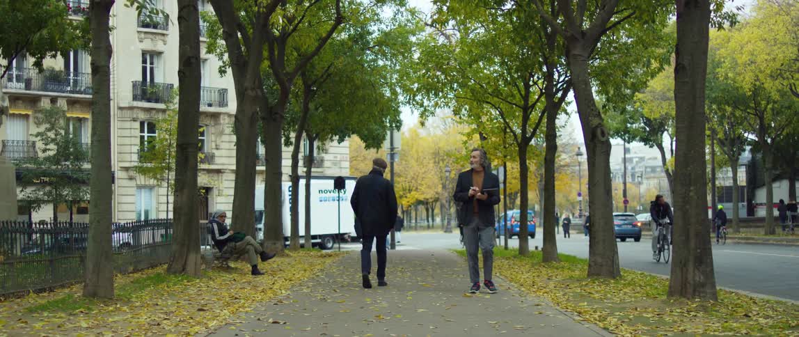 Pedestrians walk down sidewalk paralleling busy street with fallen leaves on ground and an apartment building in the background