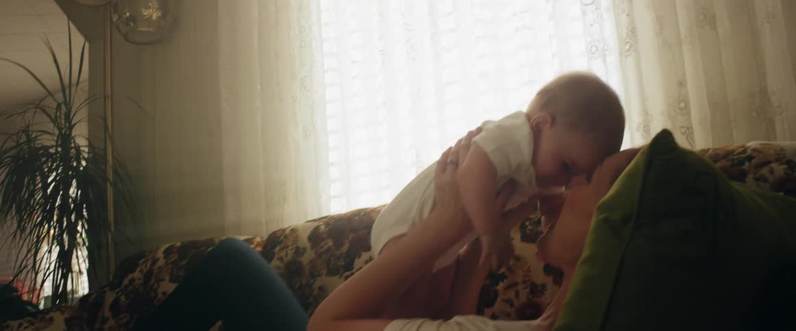 A mom lying on the couch lifting her baby up then back down smiling with sun shining through the window