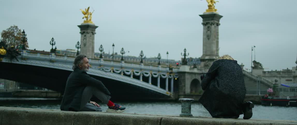 A photographer takes a photo of a man as he sits on a riverbank near downtown
