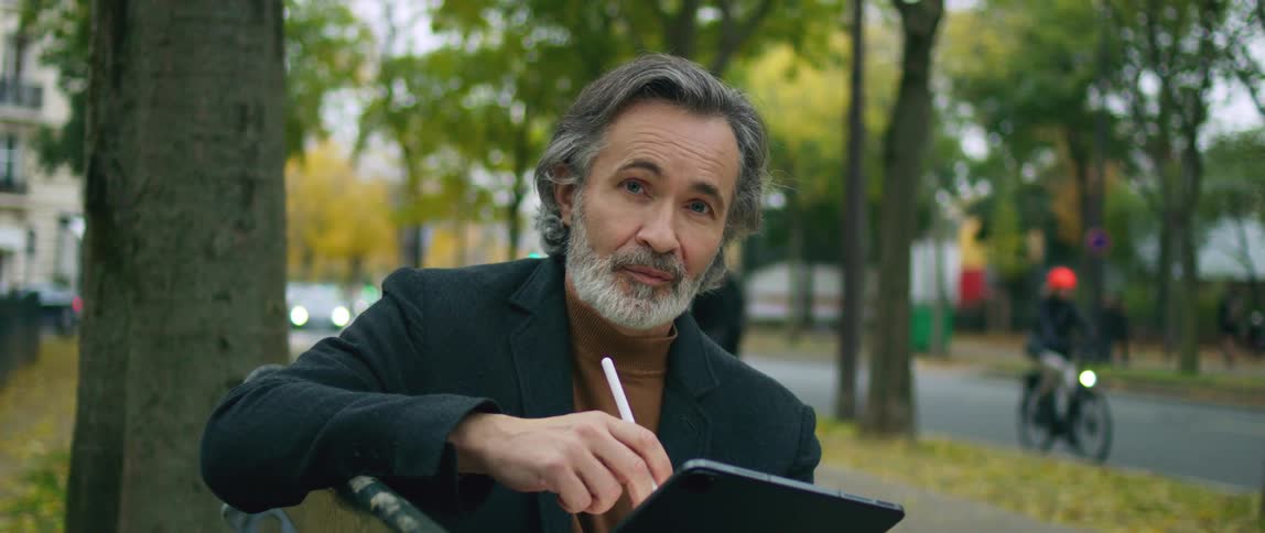 A man poses on a park bench with his tablet as a motorcyclist rides past