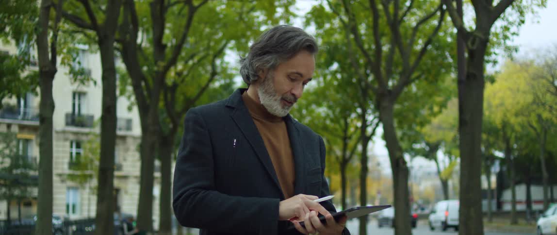 A man is standing in a park near trees using an electronic tablet as traffic passes by on the street