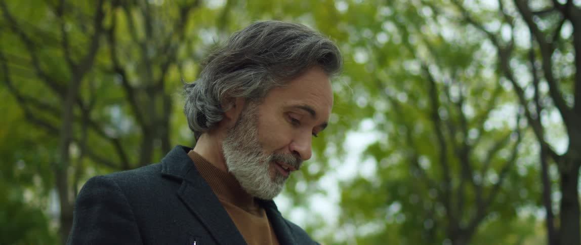 A man is standing outside among trees and he looks up and then looks down at something and smiles