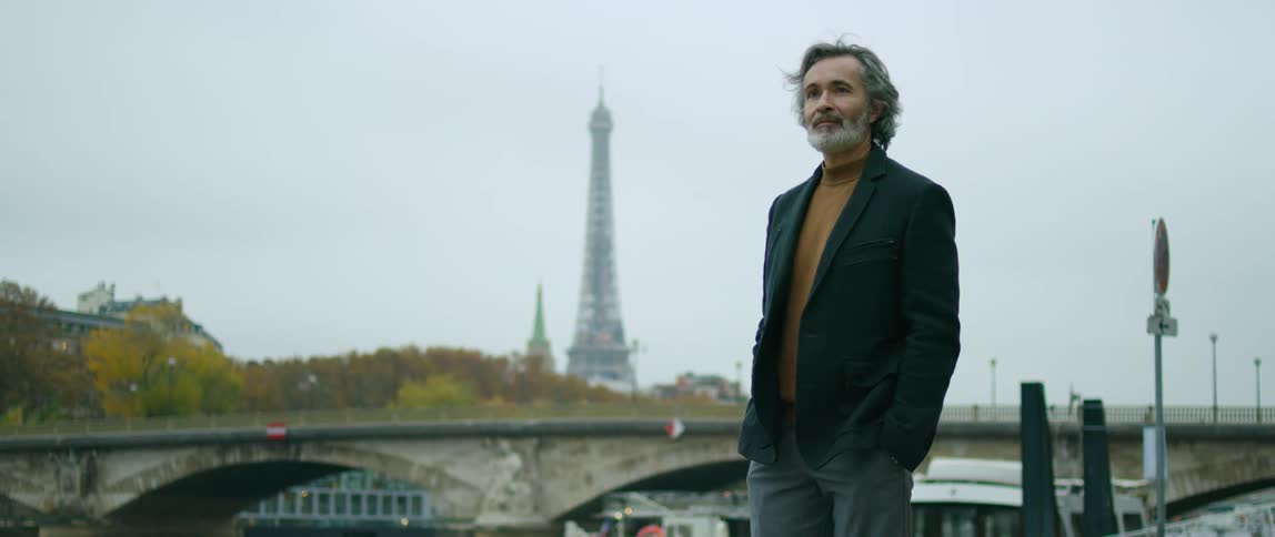A man is standing outside near a railing and a bridge and the eiffel tower is in the distance