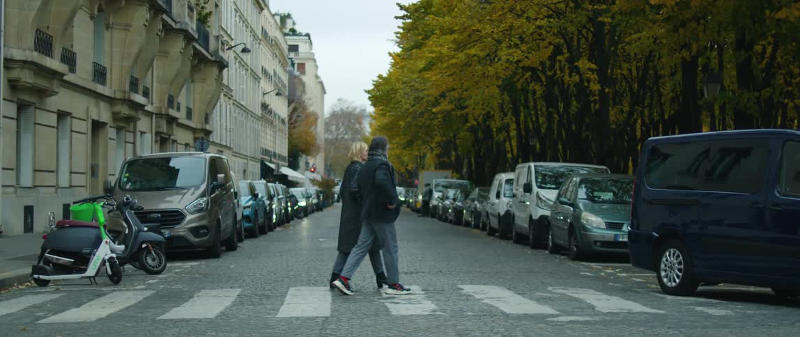 A man and woman cross a one street lined with vehicles paralleled parked on both sides