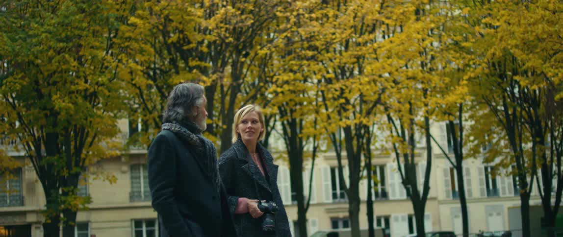 A man is walking through a park with a blond woman holding a camera and there are cars parked in front of buildings nearby