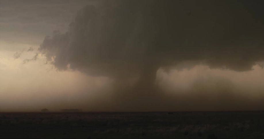 Funnel cloud moves across ground near building on the horizon
