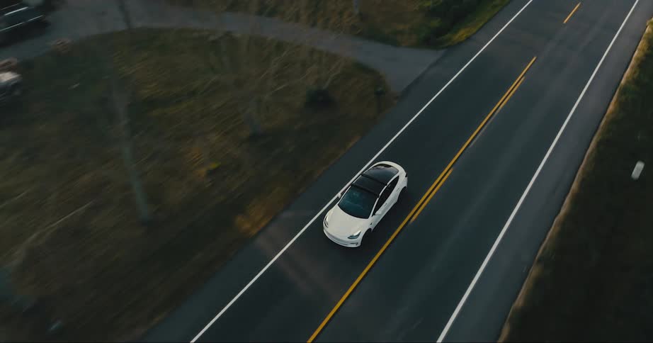 A white car travels on an empty road past yards and fields