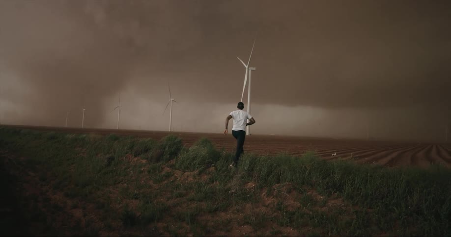 A man hops out of a vehicle on a dirt road and runs onto a field with wind turbines