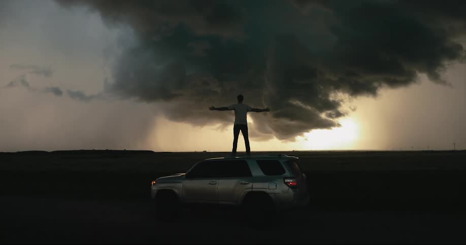 Man stands on roof of vehicle facing dark storm clouds obscuring sun