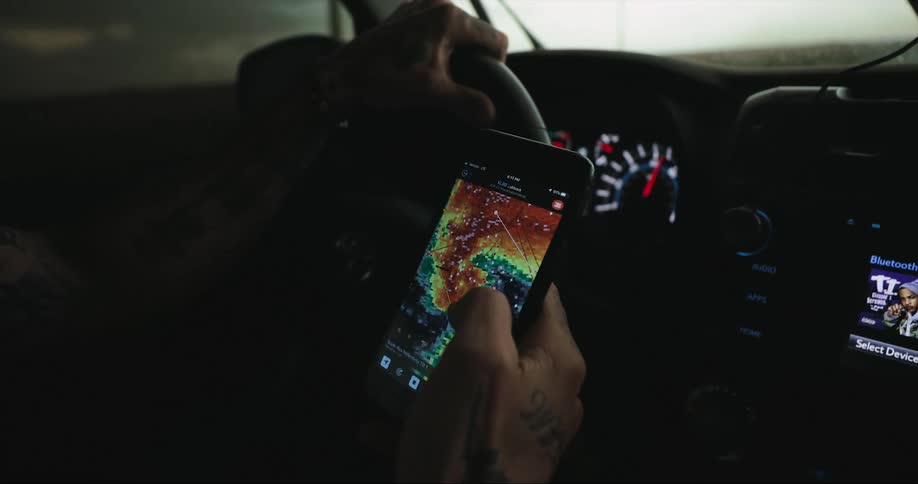 A person driving holds a smart phone in their hand as they track a storm front