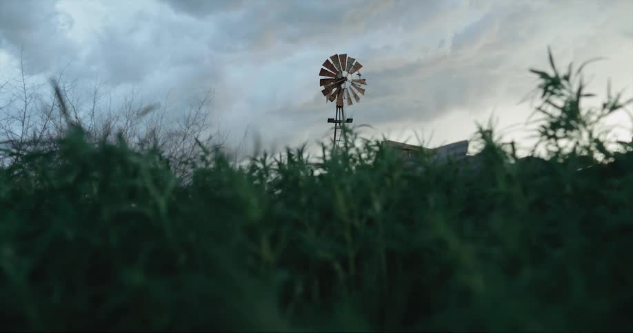 A farm with a windmill pump is surrounded by planted fields as the sky above is filled with clouds