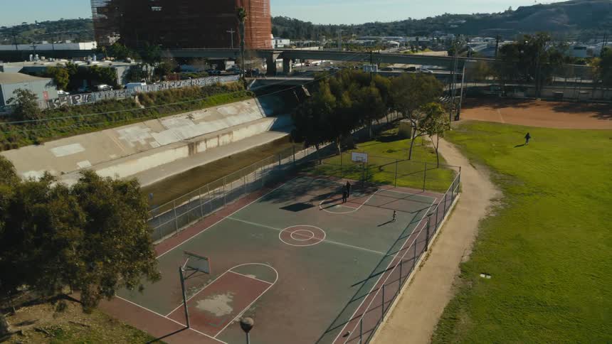Men play basketball on court at town park as child watches and another person walks across field