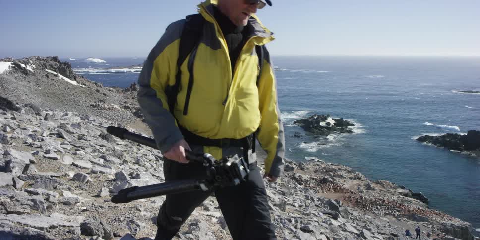 A man walks along the rocks of a shore carrying a tripod