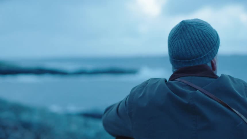 A man is standing along the shore watching the waves crash into the rocks