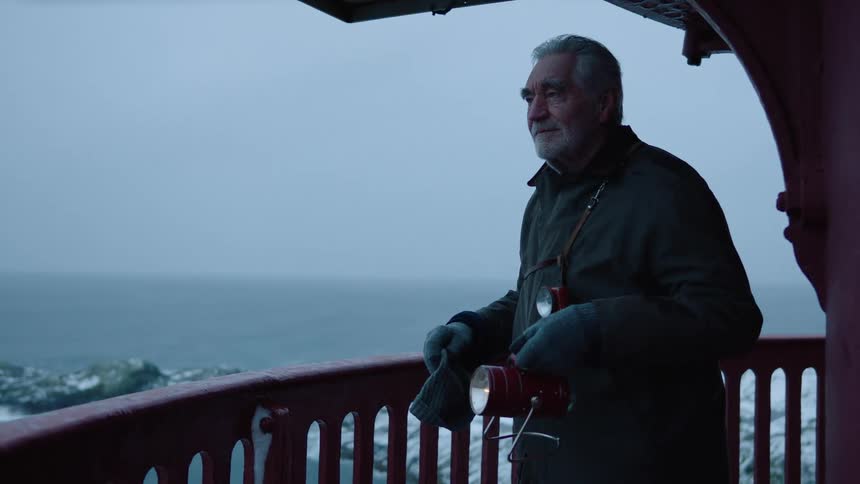 Older man stands on balcony overlooking shore holding signal light