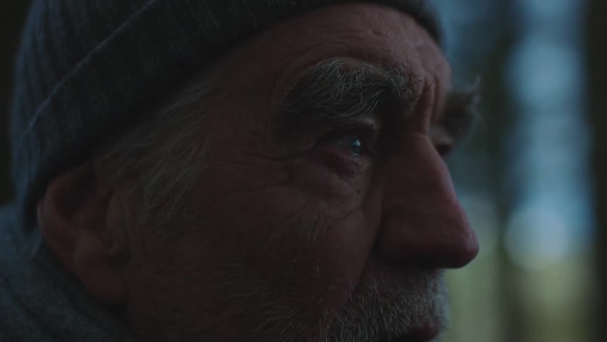 A graying man in winter clothing smokes a pipe outdoors during the day