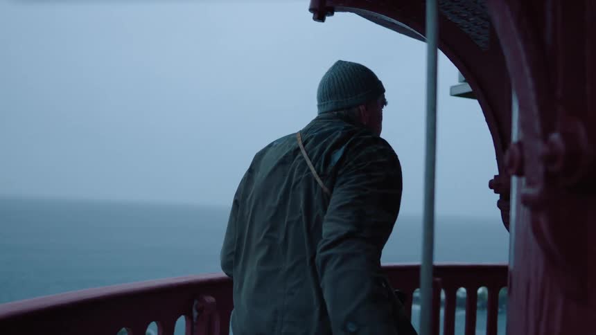 A man walks out onto a lighthouse walkway standing at the railing above the ocean waves