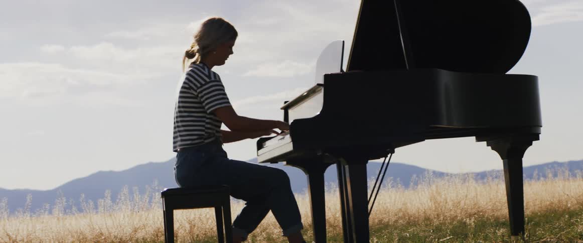 A young woman playing the piano outside with hills on the horizon and wind blowing her hair and the grass