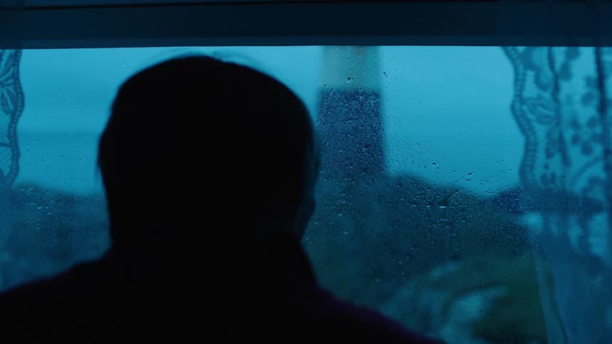 Man stands near window dotted with condensation staring out at shore and ocean