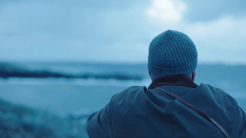 A man in a knit cap sits and looks out upon the ocean waves crashing against a rocky shoreline