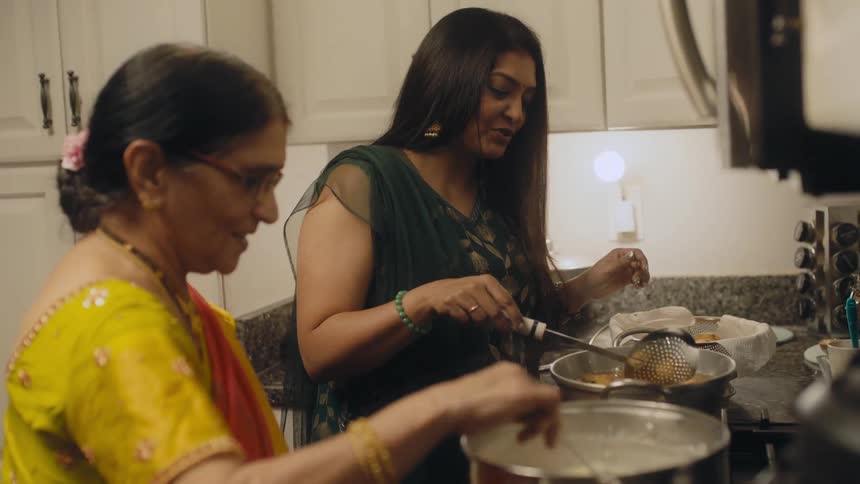Women with straight hair glasses yellow and green saris cook and fry food in the kitchen