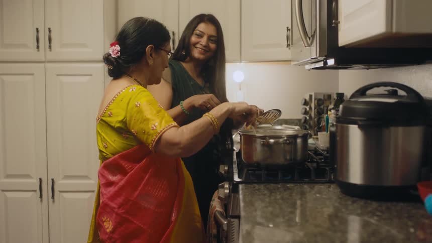 An elderly women stands at a stove and stirs the contents of a pot while a younger woman prepares food nearby