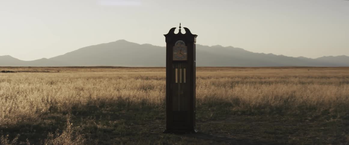 A grandfather clock sits in a field with mountains in the distance
