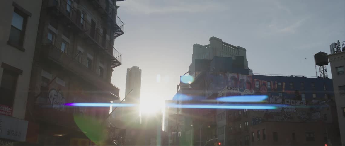 View of buildings on a downtown street as the sun shines brightly behind skyscrapers in the distance