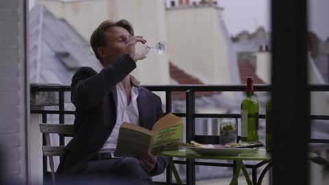 Man sitting on a balcony with a book and drinking wine