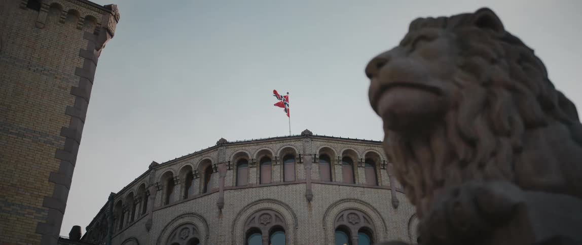 The flag of Norway billowing in the wind above the Storting Building in Oslo, Norway, with an unfocused view of a lion statue in the foreground.