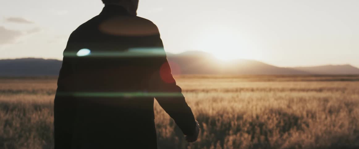 A man is walking near a field of crops as the sun shines brightly over a mountain in the distance