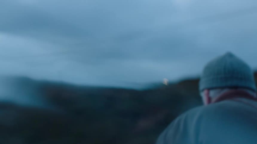 A man walking through a hilly countryside on a cloudy evening stops to look at his surroundings before continuing uphill