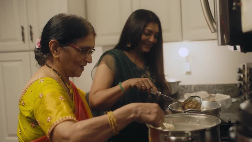 Mother and adult daughter stand in kitchen smiling as they cook and talk