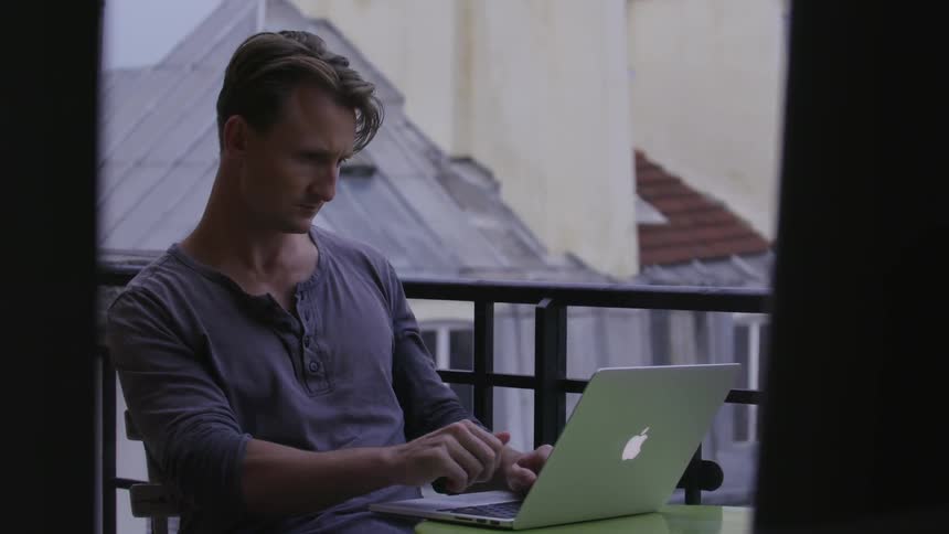 A man sits on a balcony during the day typing on a mac laptop