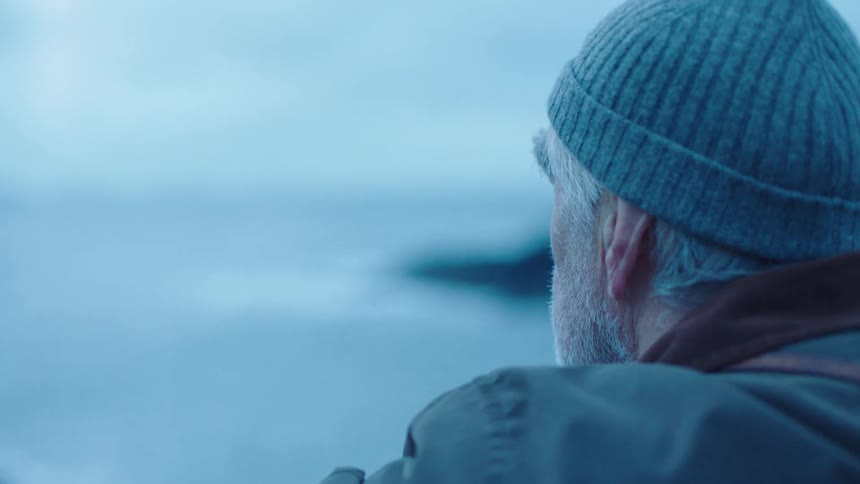 Elderly man wearing knit hat looks out to sea and around