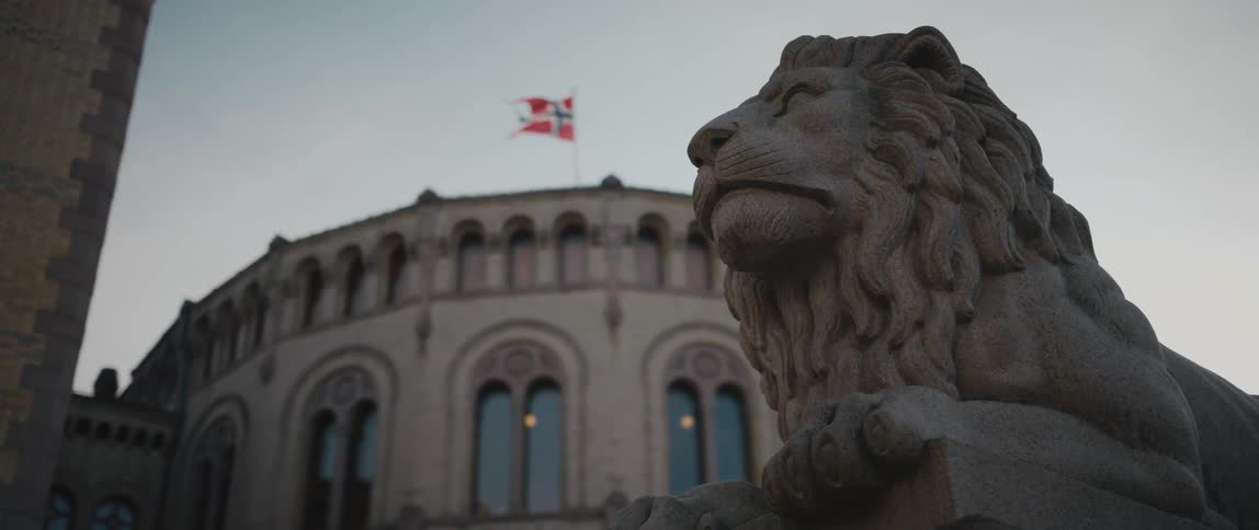 View of the Lion Statue outside of the Norwegian Parliament Storting Building in Oslo, with the flag of Norway billowing in the wind in the background.