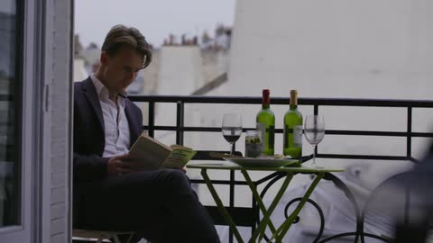 A man in a nice suit sits on a balcony during the day with a book and some wine
