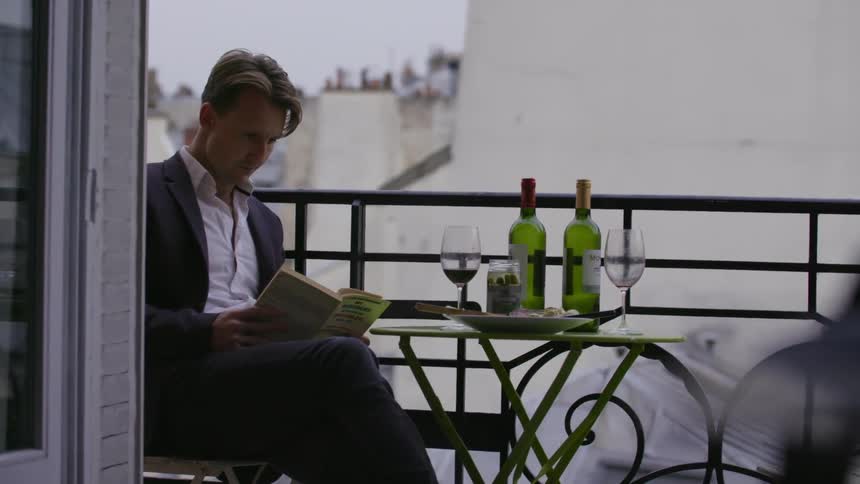 A man in a nice suit sits on a balcony during the day with a book and some wine