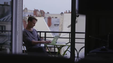 A man is sitting on a balcony using a laptop