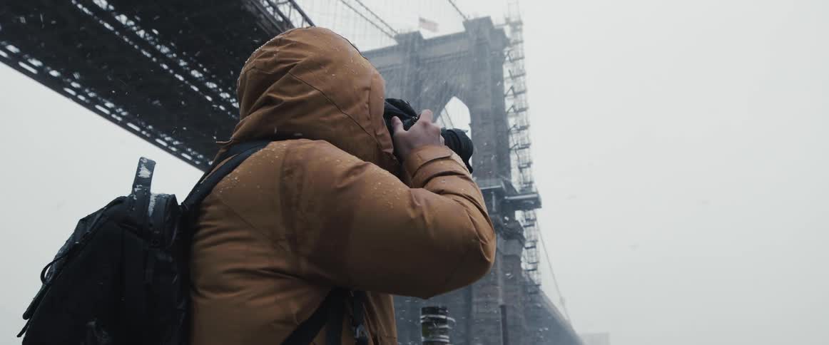 A man lifts his camera and turns to photograph a city bridge in the snow