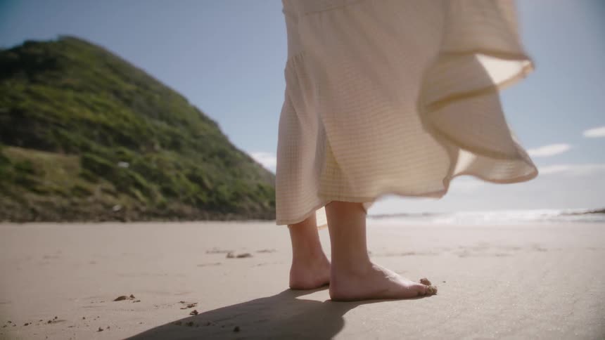 A barefoot woman in a dress stands on a sandy beach on a sunny day