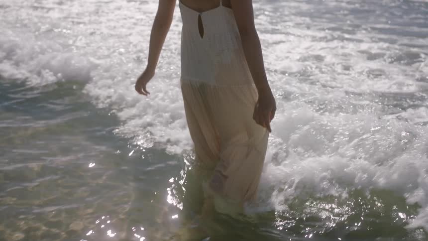 A woman wearing a sundress is on a beach walking into the water as waves roll in