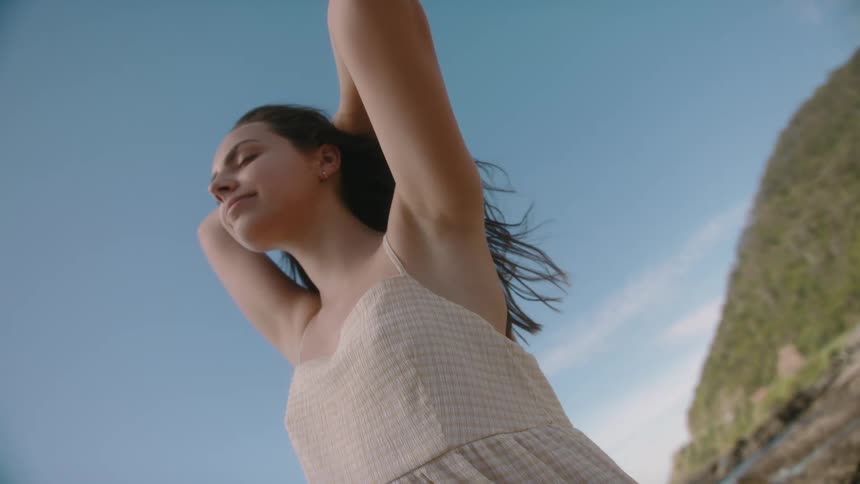 A young woman stretches while standing outdoors on the coast on a sunny day