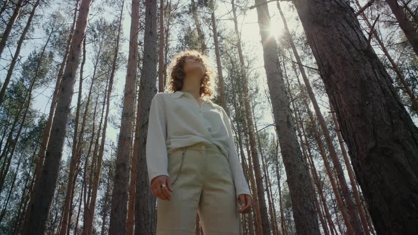 A woman standing in the woods lifts her head and stares up