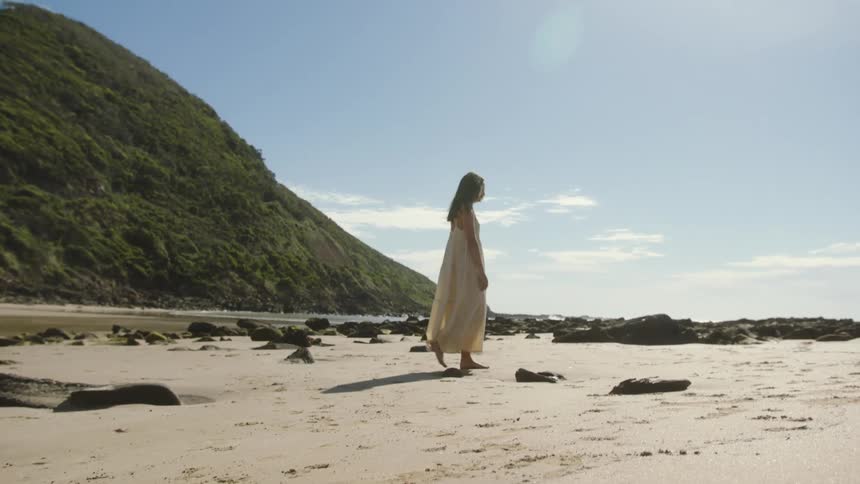 A woman in a dress walks barefoot along a rocky beach on a sunny day