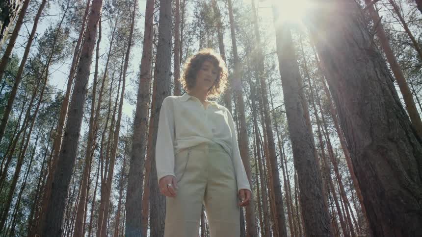 A woman standing in a forest looks down at the ground as the sun shines down