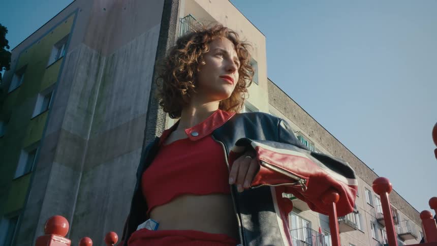 A woman is standing on a street leaning against a red fence and looking around and there is a building behind her