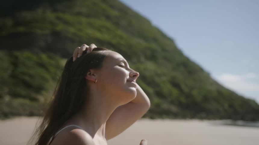 A woman runs her hands through her hair while walking along a beach on a sunny day