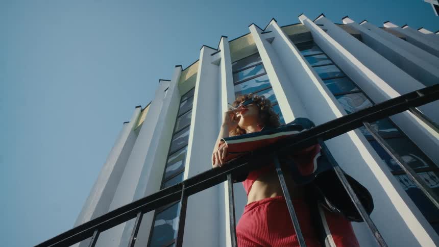 A woman wearing red pants and a jacket is leaning against a rail smoking and there is a tall building behind her