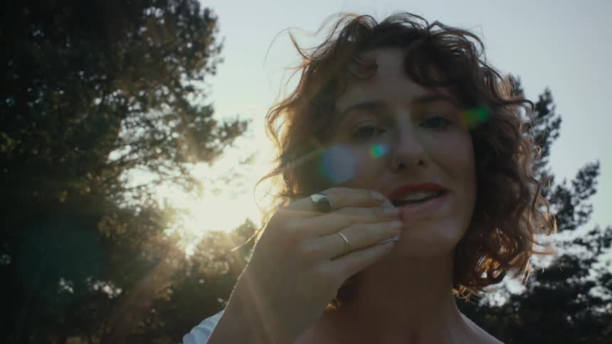 White woman standing outdoors in front of some trees taking the lipstick out of her mouth